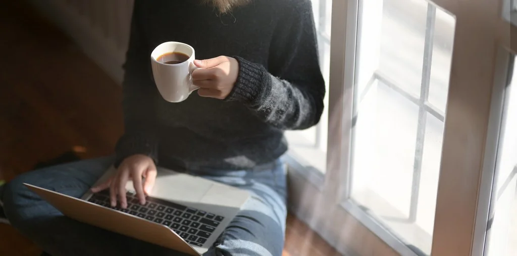 Person typing on a laptop while holding a cup of coffee by a window