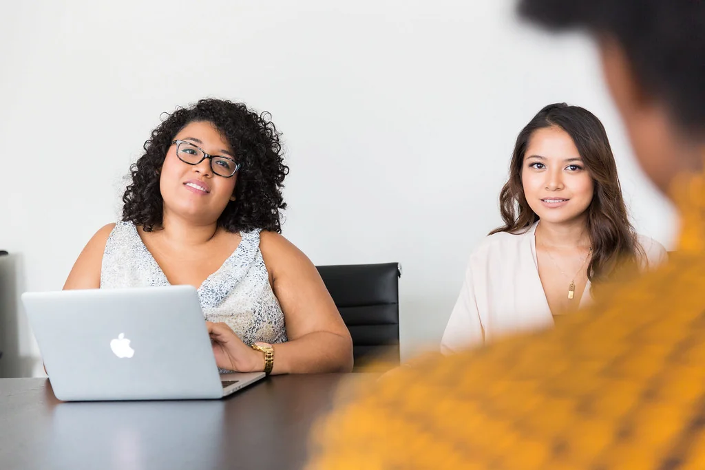 Two women at a meeting table, one typing on a laptop